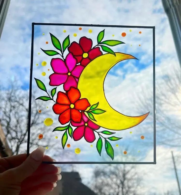 A glass painting of a yellow crescent moon and pink flowers held against the sky
