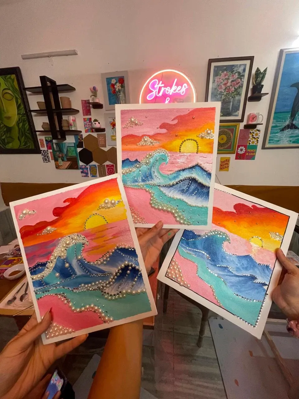 Three pearl art seascapes held up in front of the lit Strokes & Verses neon sign